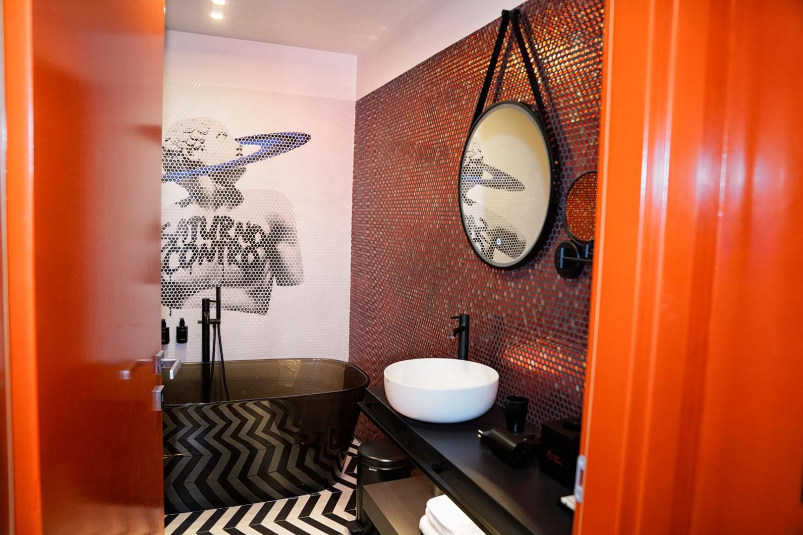 Bagno con vasca nera, murale Saturno Contro in mosaico, specchio tondo e lavabo