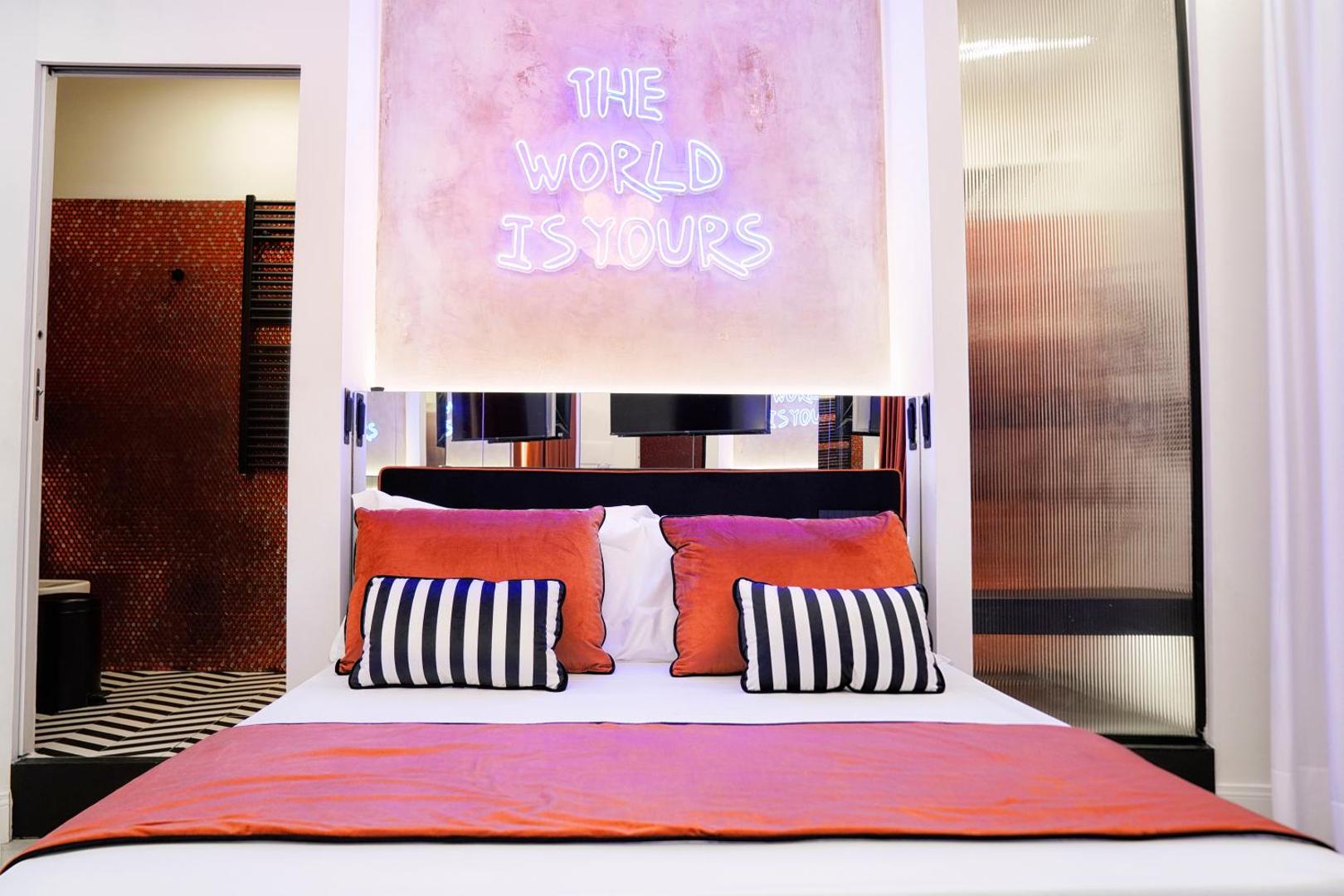 Letto matrimoniale con insegna al neon The World Is Yours e cuscini arancio e a righe